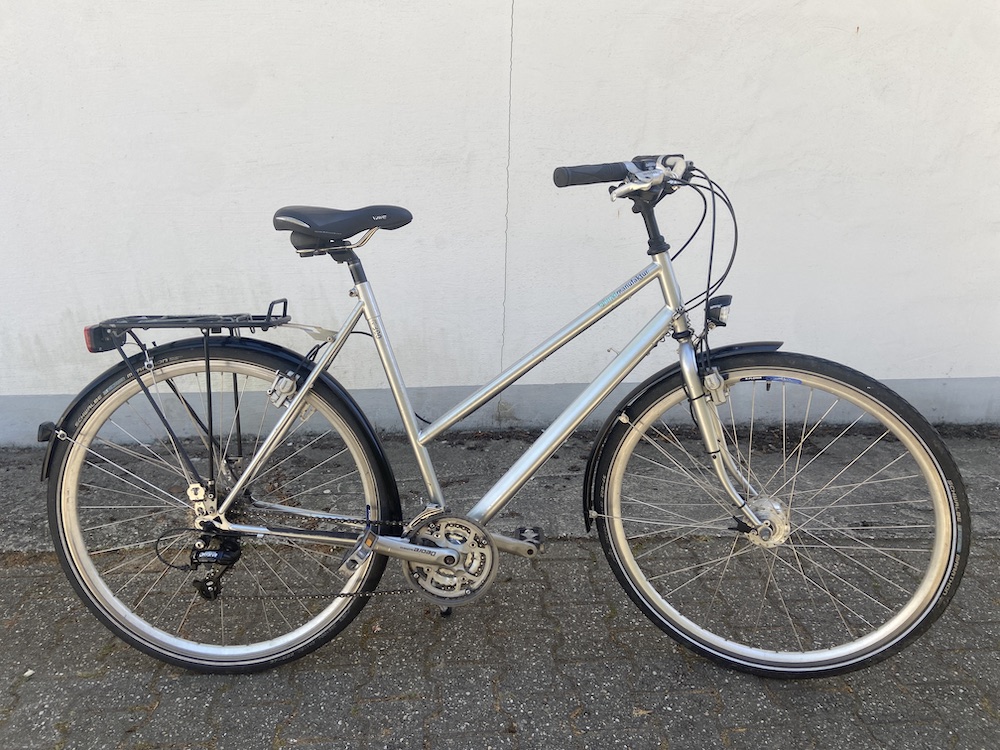 Silbernes Trekkingfahrrad mit Gepäckträger, Satteltasche und Schutzblechen vor grauer Wand und Boden.
Verkauf gebrauchter Fahrräder Aachen