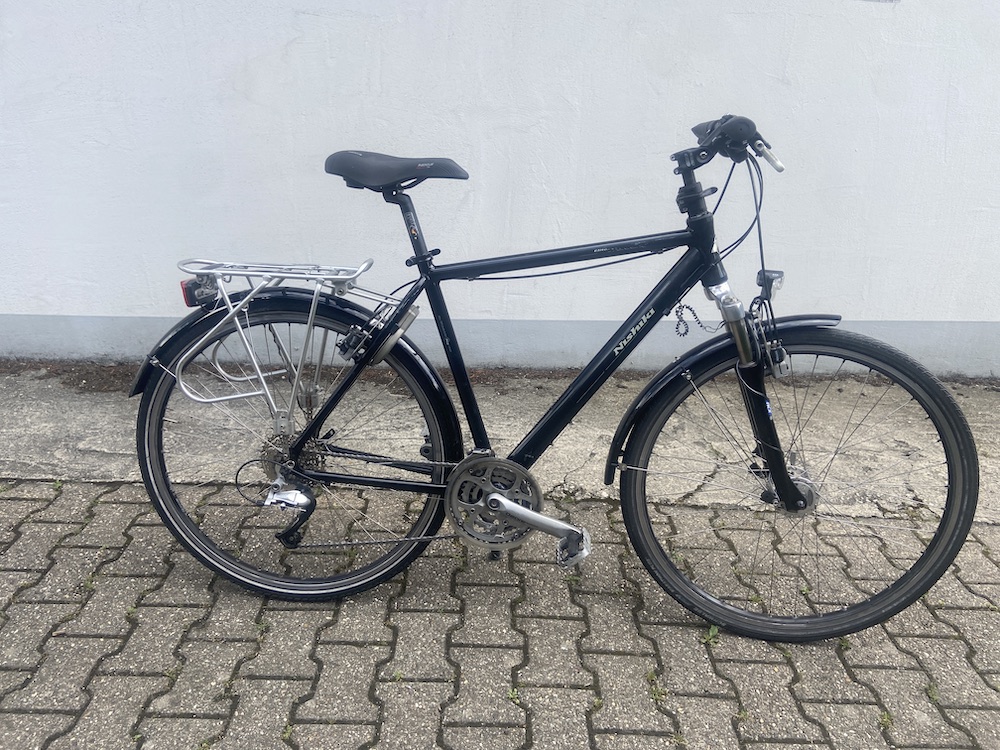 Schwarzes City-Fahrrad mit Gepäckträger, Schutzblechen und Sattel vor weißer Wand auf gepflastertem Boden.
Verkauf gebrauchter Fahrräder Aachen